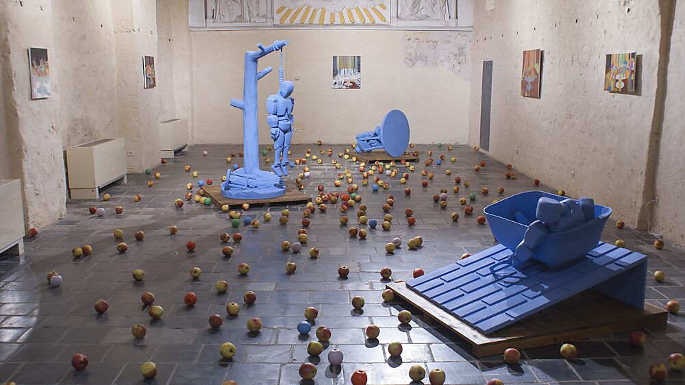 ‘Strange Fruit’: Tentoonstellingszicht met meerdere blauwe robotfiguren op platforms in een kapelruimte; vloer bezaaid met echte appels (sommige gekleurd), schilderijen aan de muren, Gasthuiskapel, Borgloon, 2015.