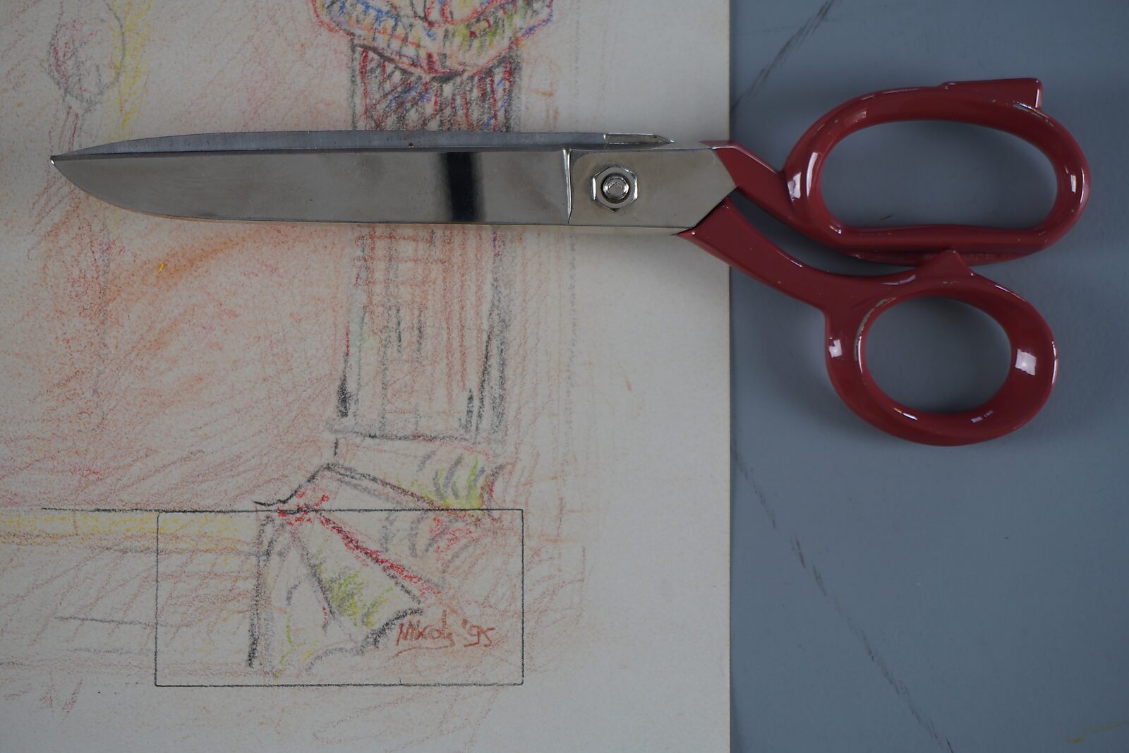 Schaar met rode handgreep ligt op een tekening op papier; een rechthoekige uitsnede markeert een fragment met signatuur en kleuraccenten.