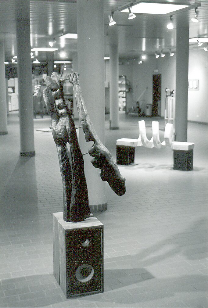 ‘Houten spiralen en metalen’: Exhibition view with metal sculpture of a standing figure on a pedestal; abstract forms in an open exhibition space, black-and-white photograph, CC Leopoldsburg, 1999.