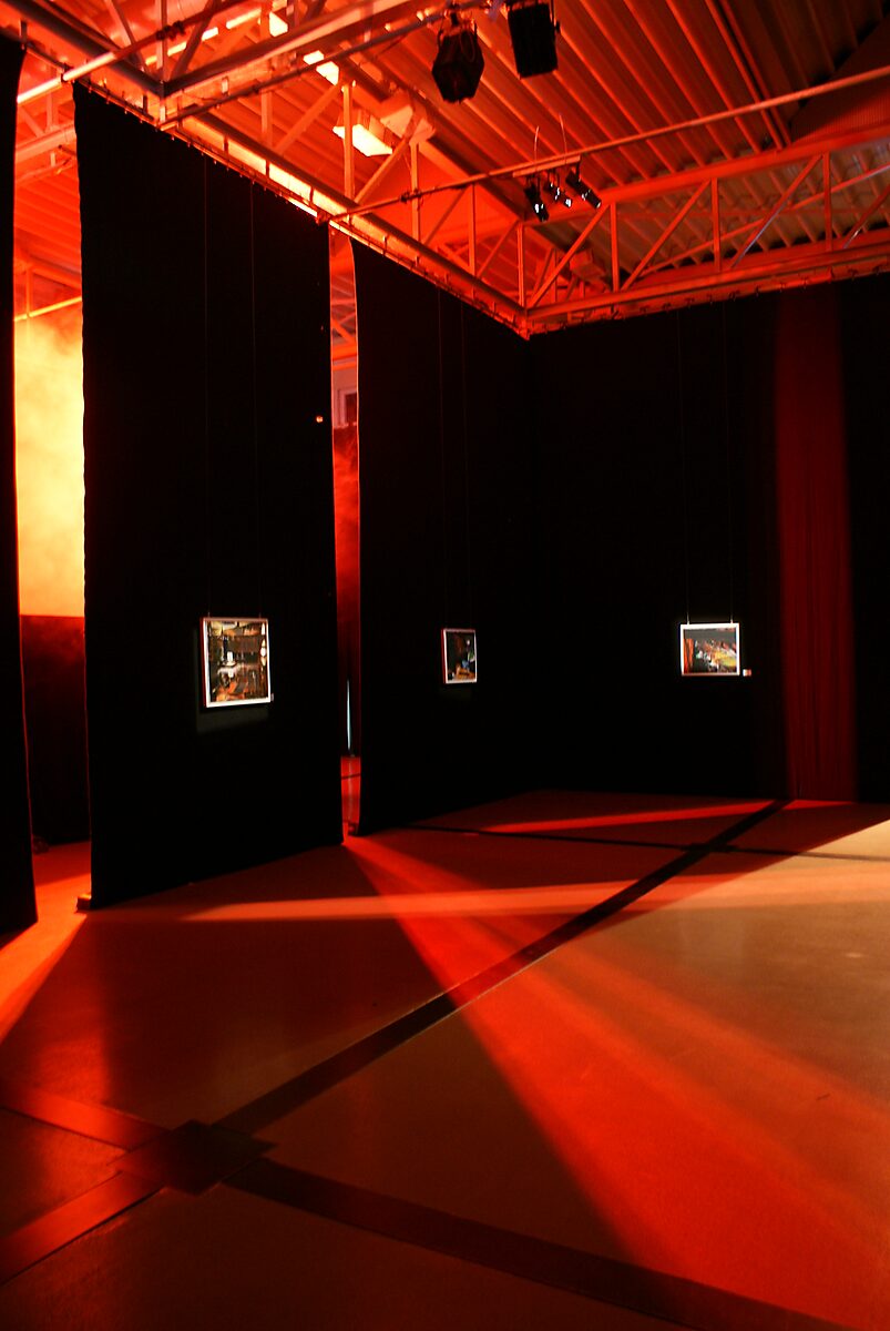 The Modern Cavemen — paintings presented within an angular arrangement of curtains; red light cuts diagonal beams across floor and walls, activating the space as part of the work.