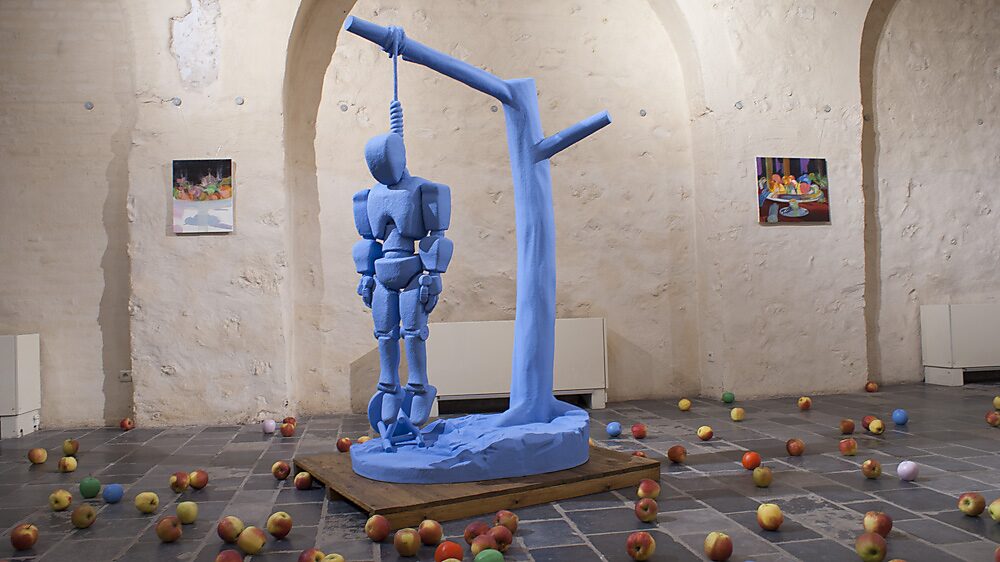 Strange Fruit — exhibition view at Gasthuiskapel Borgloon showing a suspended robot figure beneath a tree-like structure, surrounded by scattered real and painted apples activating the sculptural environment.