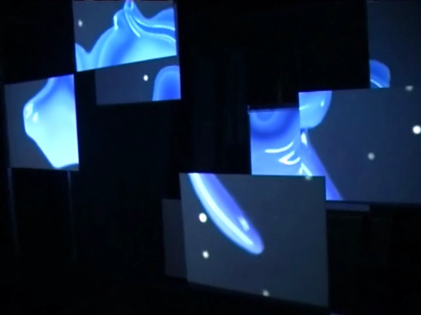 Vloeibare vormen doorheen de ruimte — videostill van de installatie waarin blauwe vloeibare vormen over de panelen glijden, gefragmenteerd door de opstelling en wegvloeiend in de donkere ruimte.