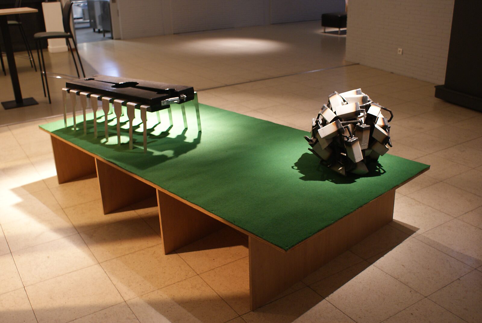 De moderne holbewoners — extra opstelling met de sculpturen Pilory Chip en Camera bal op een lage tafel met groen oppervlak; de objecten verwijzen naar observatie, controle en technologische surveillance binnen de tentoonstellingsruimte.