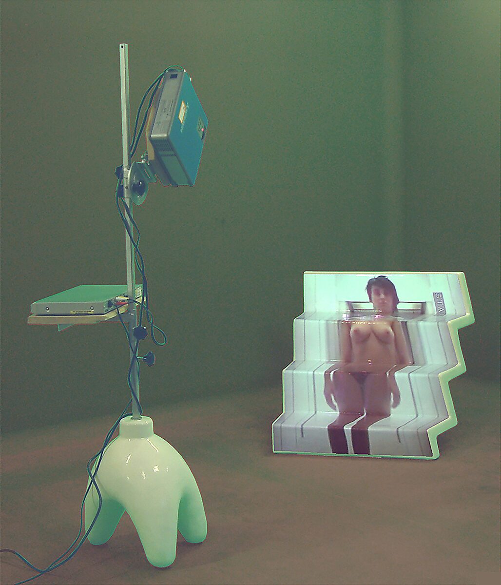 Nu descendant par un ascenseur — edited photograph of the installation featuring a video projector and luminous stepped sculpture in which a female figure appears as a projection, emphasizing the relationship between body, image, and technology.