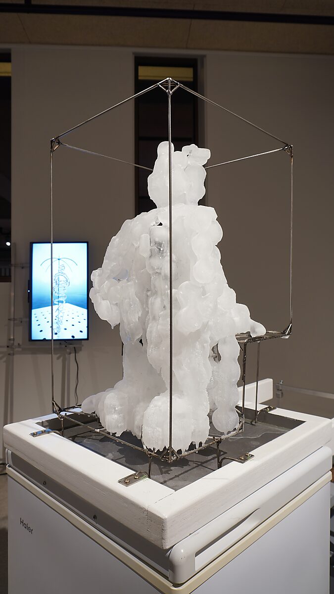 The Icy Man — the final ice sculpture inside a metal frame on a freezing installation; in the background a screen shows the 3D animation of the virtual version of the figure.