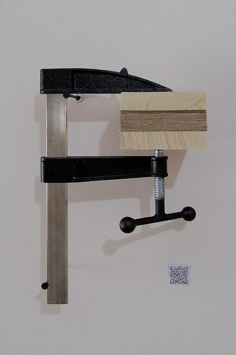 Schilderijen.zip — een samengeperst schilderijblok wordt tussen twee houten platen vastgezet in een metalen lijmklem die verticaal aan de muur is bevestigd; de gelaagde snijranden van het doek vormen een compacte horizontale stapel.