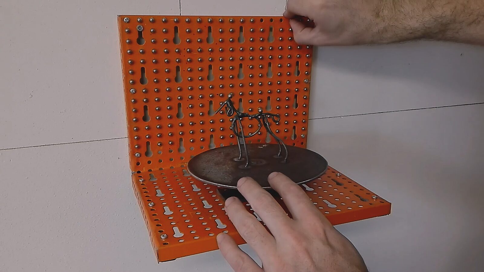 De draaihond — videostills: ijzerdraad hond op een ronde metalen schijf, geplaatst voor een oranje geperforeerde wand; handen brengen metalen bolletjes aan.