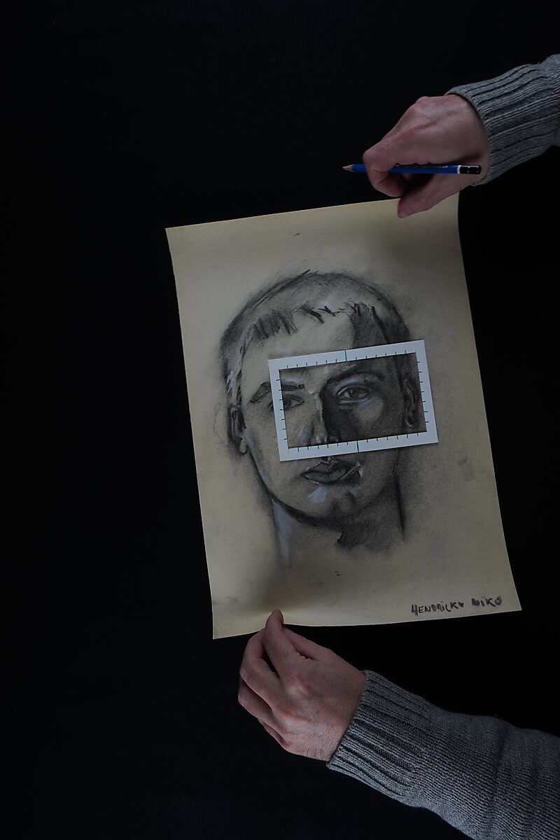 Benoemens-waardvlinders — Naming–Value Butterflies — proces tekening: studie zelfportret 2 PHIKO (1993), Gevangen (2025) — een houtskool zelfportret wordt vastgehouden terwijl een rechthoekig kader met schaalverdeling over de ogen ligt.