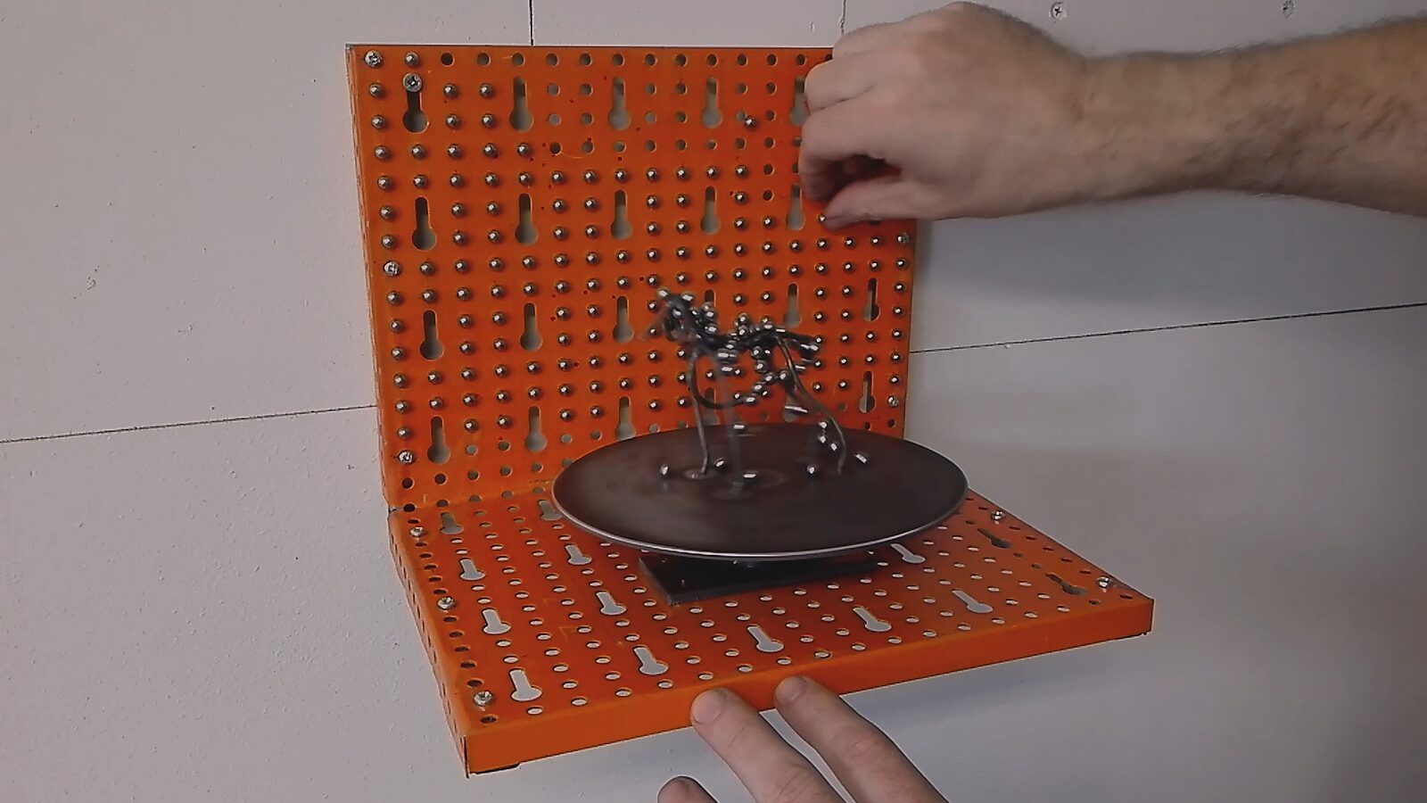 De draaihond — videostills: ijzerdraad hond gedeeltelijk bedekt met metalen bolletjes terwijl een hand nieuwe bolletjes plaatst op de vorm.
