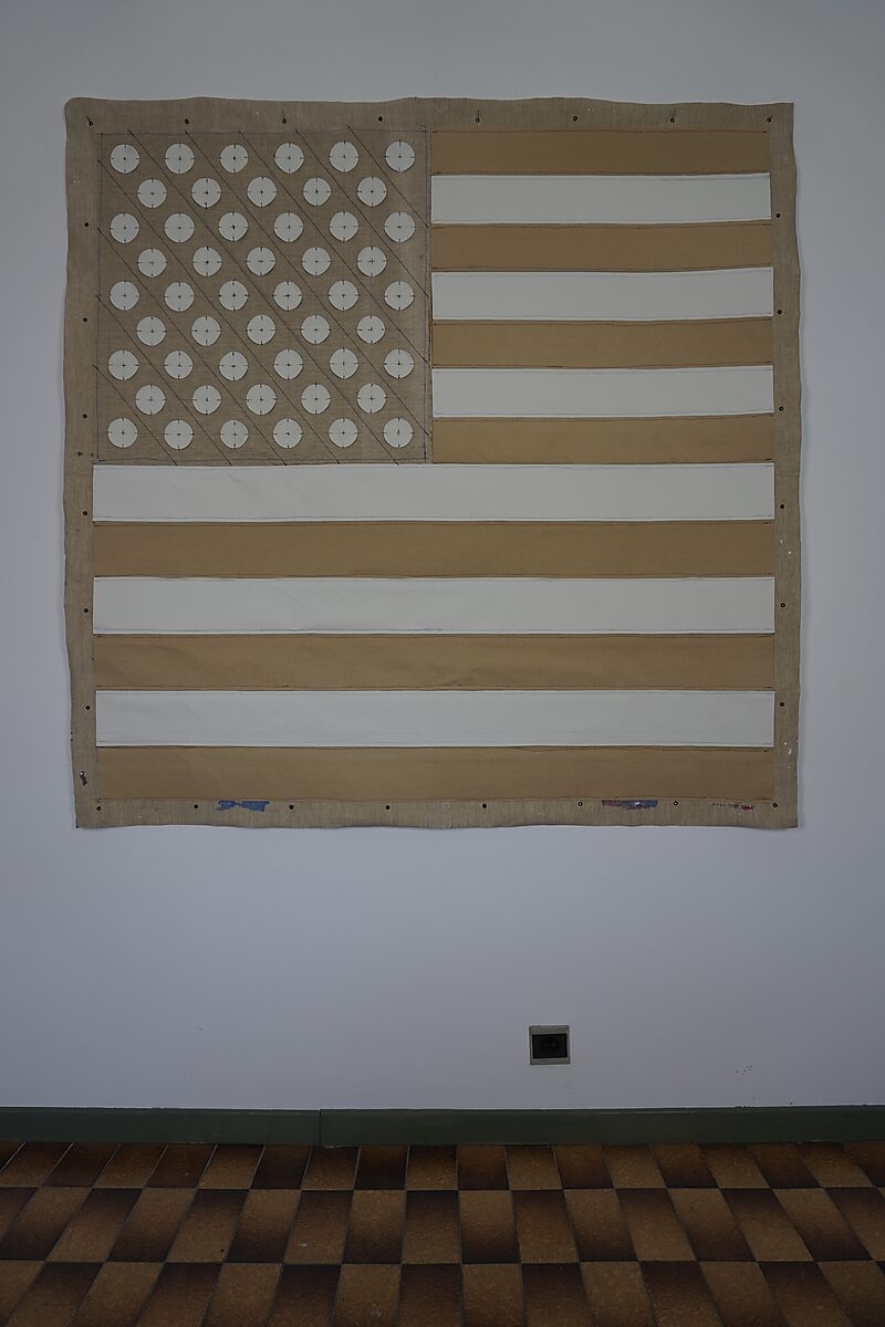Linnen doek als Amerikaanse vlaginterpretatie met horizontale stroken en een veld van cirkels linksboven, strak gepresenteerd tegen een witte muur; de tekst “MAGA” bevindt zich op de kleurzijde.