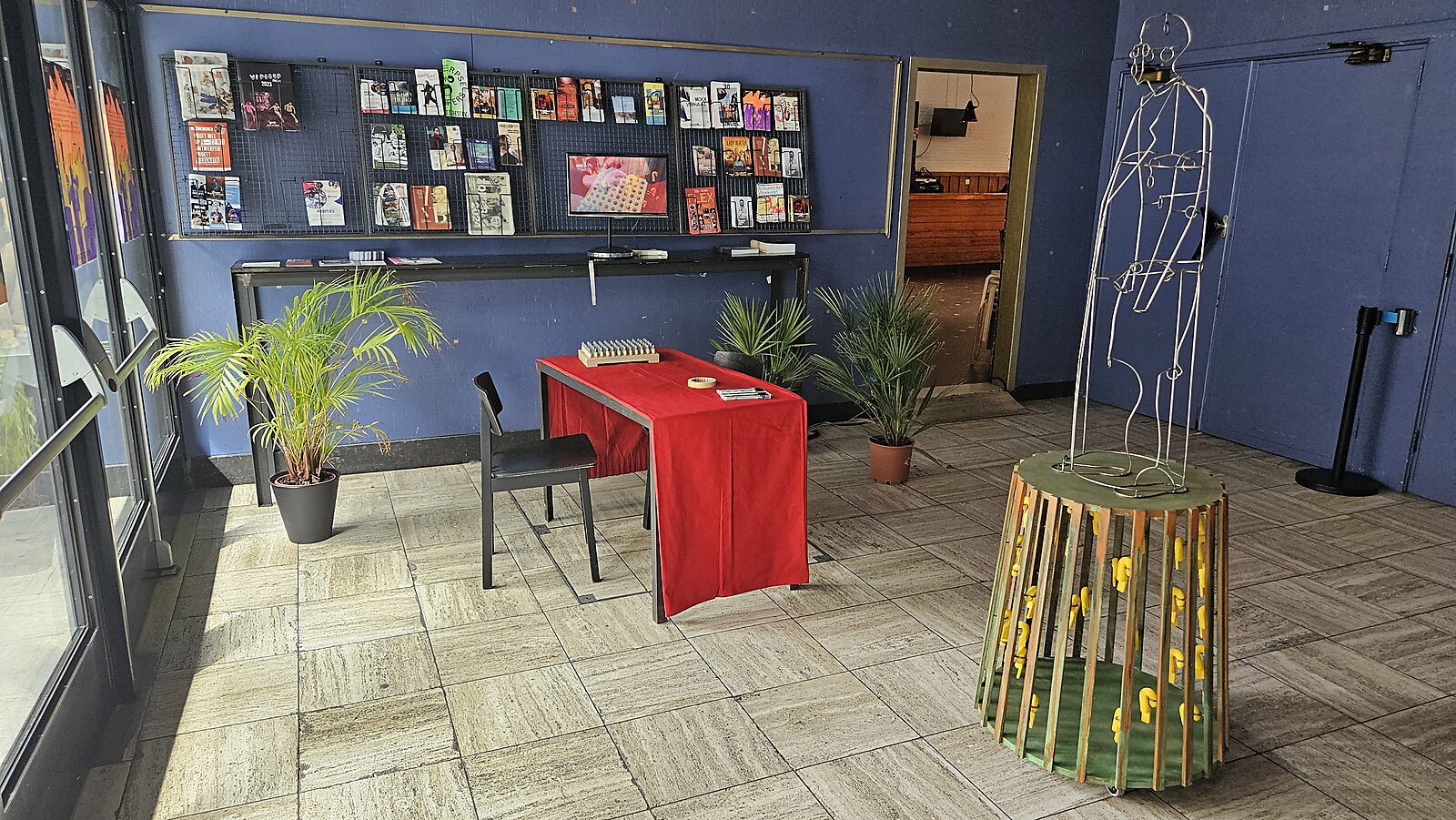 'Beyond the Black Box' Exhibition view with table covered in red cloth and chair; metal figure on a round pedestal and wall with publications in the background, Monty, Antwerpen, 2024.