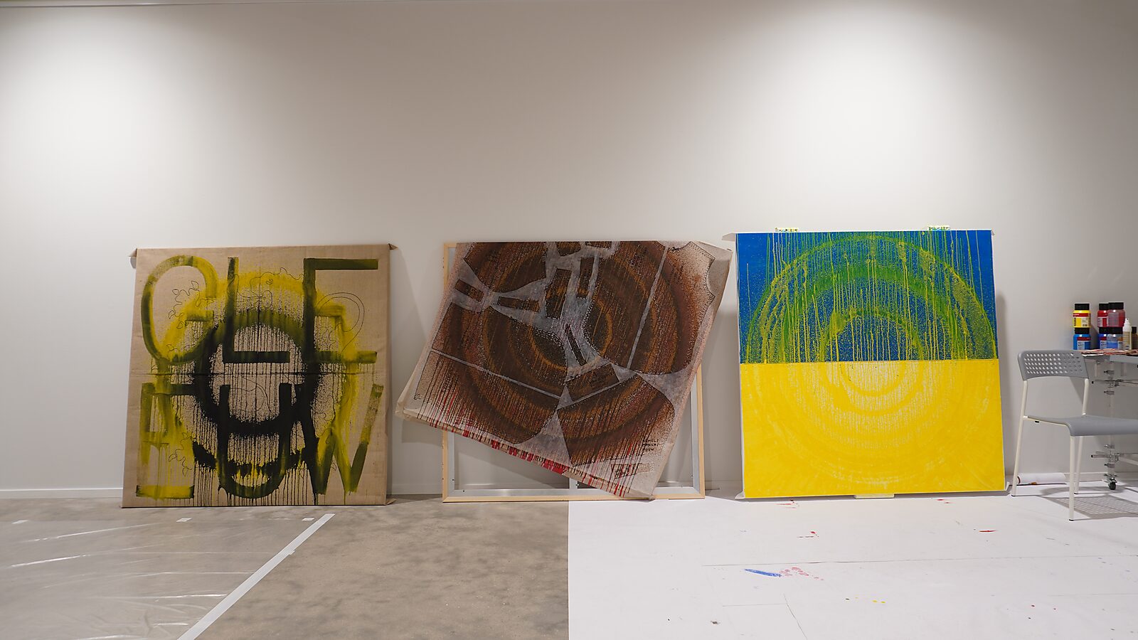 Three large canvases against the wall: on the left a green work with a white grid structure, in the center a brown layered canvas in a frame, and on the right a blue-yellow painting with circular traces; paint and materials are scattered across the floor.