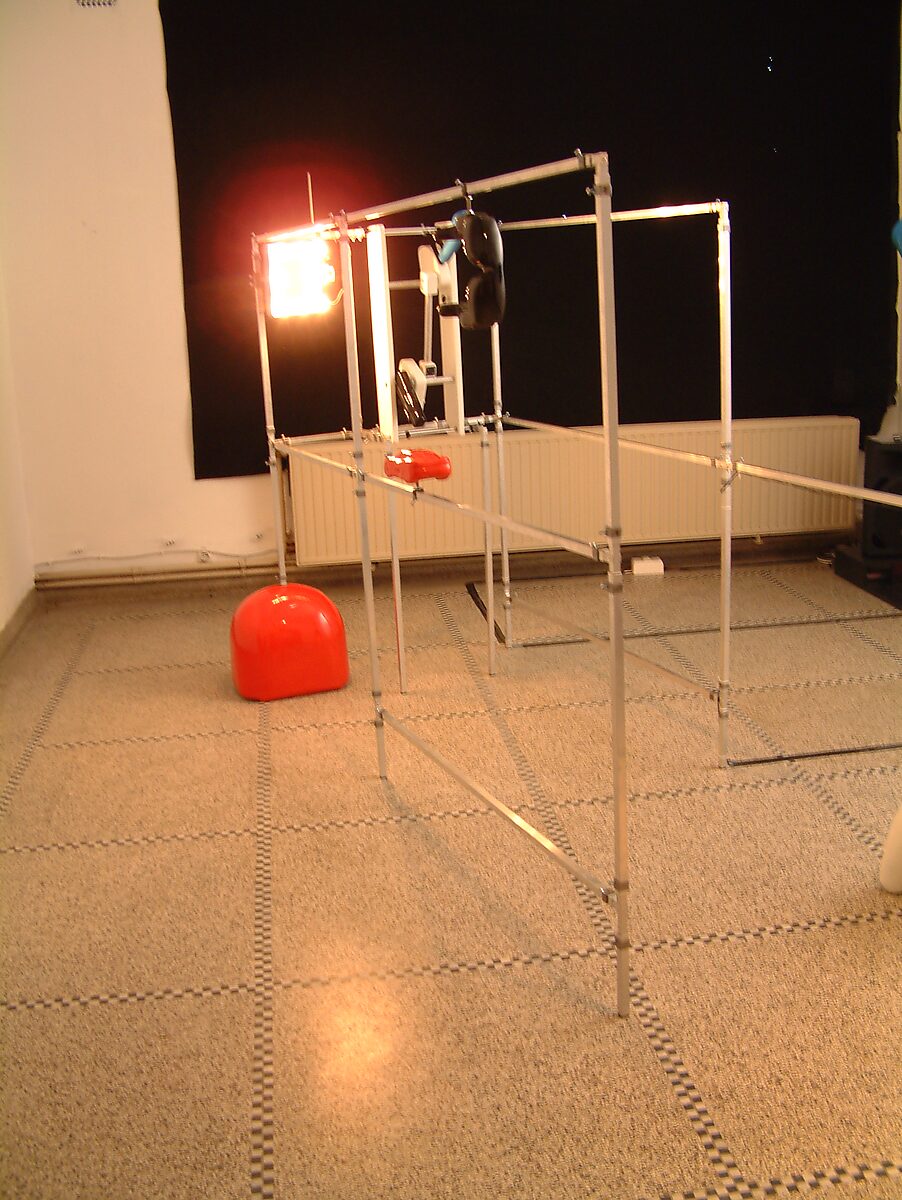 Kies en verander, Installatie voor een egoïstische rapper — foto van de installatie: ruimtelijke opstelling met metalen frames, rode objecten en een fel lichtpaneel dat de installatie verlicht.