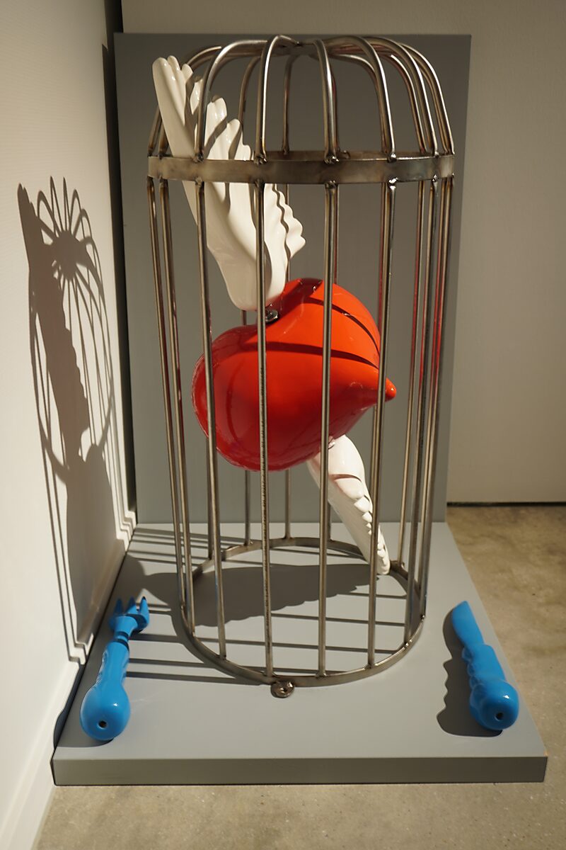 Kies en verander — Transformatiestudio nr. 1: metalen kooi met een rood object en witte handen, geplaatst op een sokkel met blauwe elementen ernaast.