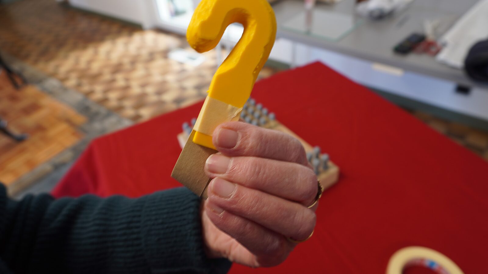 Sfincterman No.0.0 — action videostill: the yellow hook-shaped object with the attached paper strip is held up, with tape visible around the connection.