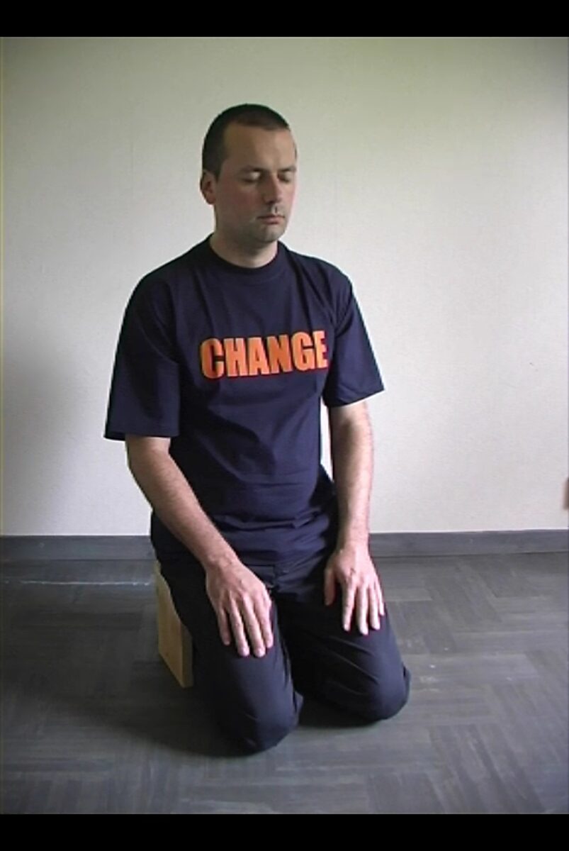 Kies en verander nr. 2 — videostill van de kunstenaar knielend op de vloer, met gesloten ogen en het woord “CHANGE” op het T-shirt.
