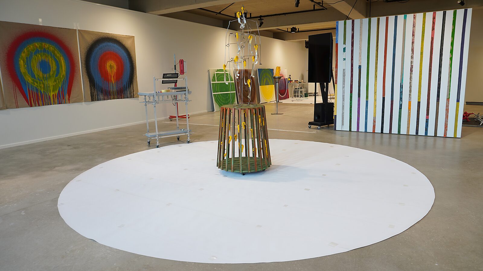 Sfincterman met kleurententakels — tentoonstellingszicht: de sculptuur staat centraal op een groot wit cirkelvlak; vanuit de metalen structuur lopen transparante en gekleurde buizen als tentakels naar buiten over de vloer, omringd door schilderijen en ins