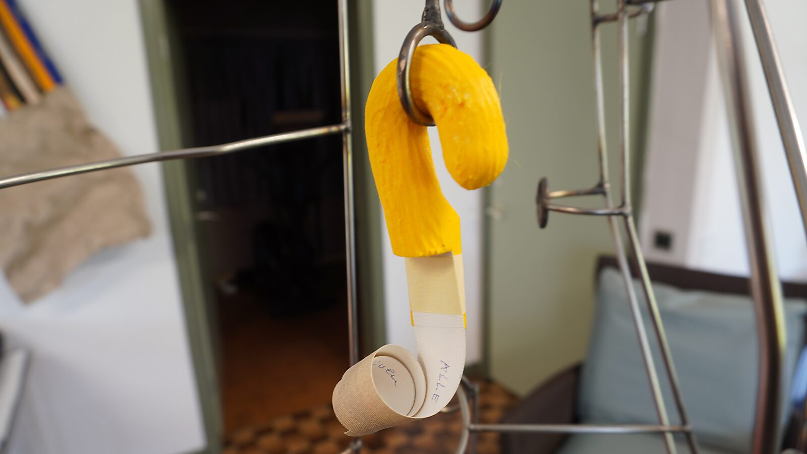 Sfincterman nr.0.0 — videostill handeling: het gele haakvormige object hangt zelfstandig aan een metalen ring, met een opgerolde papieren strook die naar beneden hangt.