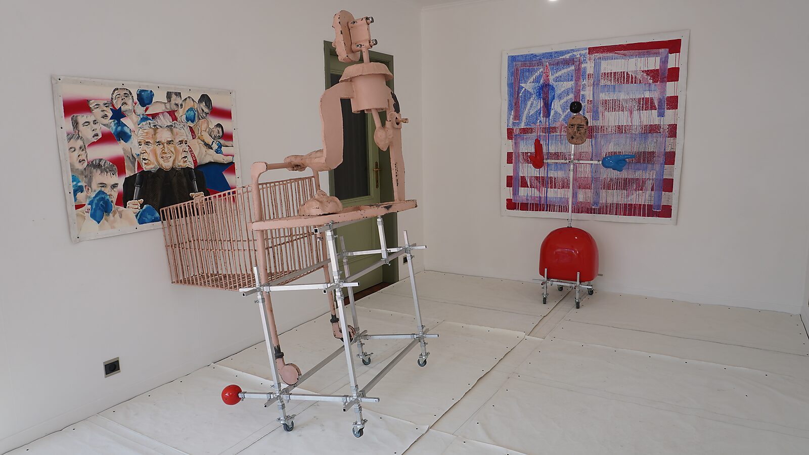 Sculptural installation on wheels with a pink figure-like object in an aluminum frame; on the wall behind hangs a canvas based on the American flag with the text “MAGA”.