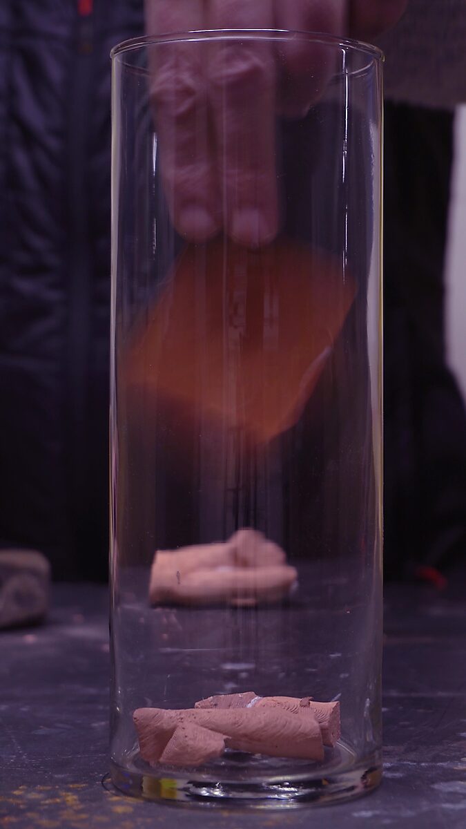 Gebroken volume — videostill waarin de eerste brokstukken van het beeldje onderin de glazen cilinder liggen.