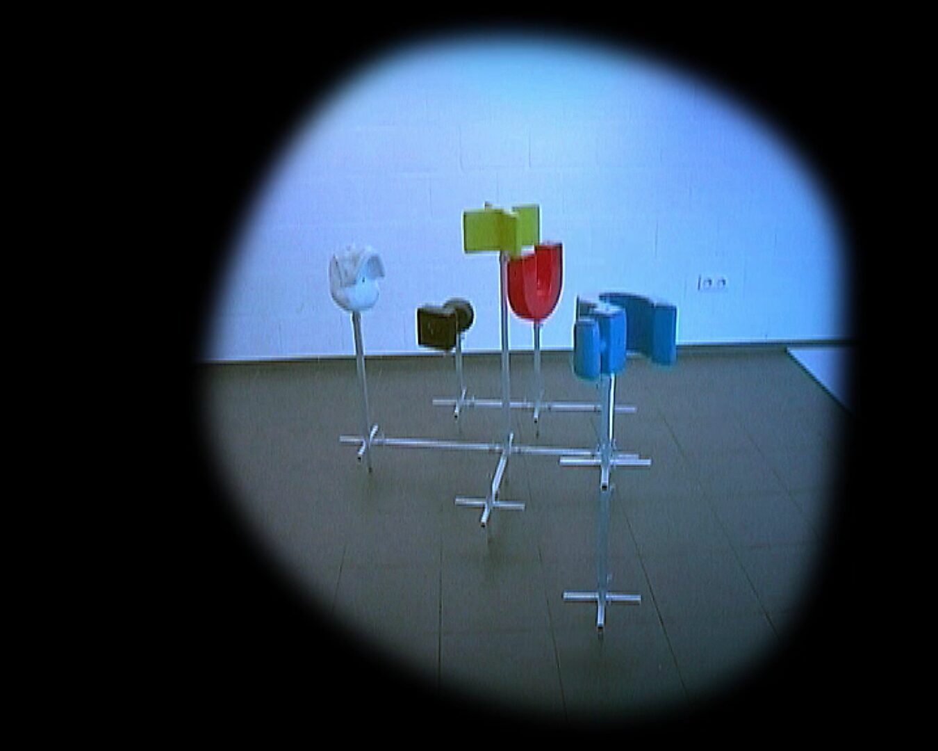 Kies en verander, Installatie in beweging — videostill: opstelling van gekleurde objecten op metalen stangen, centraal gegroepeerd in een lege ruimte, gezien door een ronde vignette.