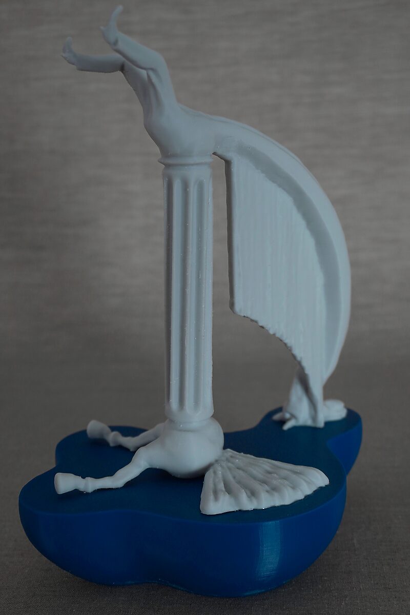 De zuilen van Ovidius, Ocyrhoë — zijaanzicht van een witte 3D-geprinte figuur die over een klassieke zuil hangt, met uitgespreide benen en een neerhangende vleugelvorm op blauwe sokkel.
