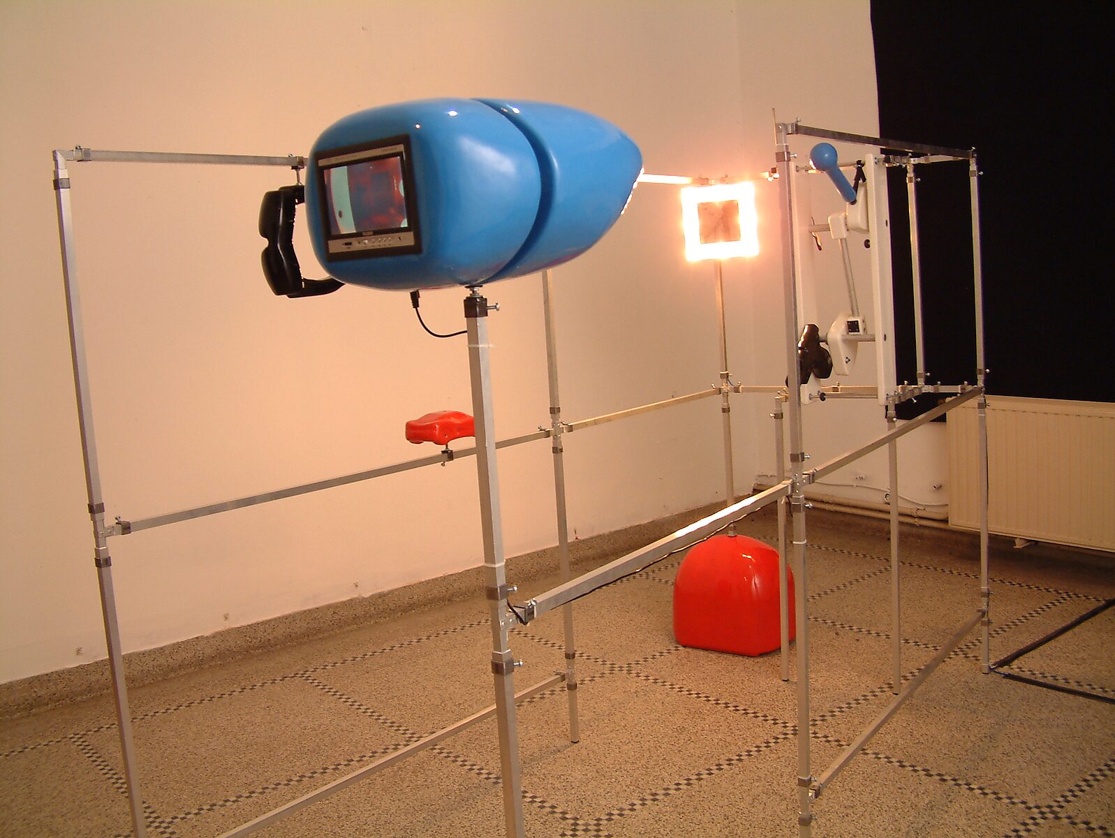 Kies en verander, Installatie voor een egoïstische rapper — detailfoto: blauw monitorachtig object met ingebouwd scherm waarop een live videobeeld van de installatie te zien is, met op de achtergrond een verlicht paneel.