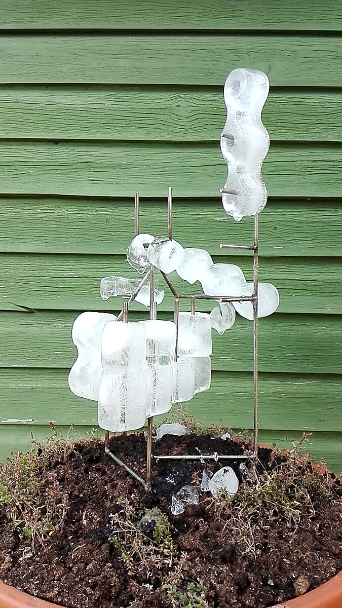 Entropic Sculpture No. 1 — videostill of the sculpture in summer with partially melted ice and loose pieces of ice on the soil around the metal armature.