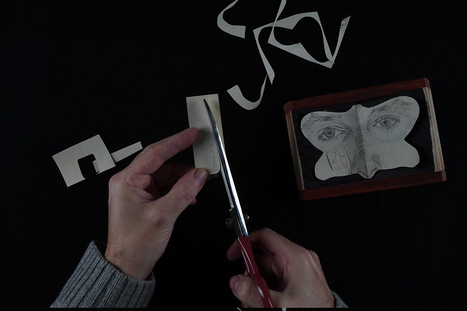 Naming–Value Butterflies — process drawing: study self-portrait 1 PHIKO (1993) — a hand cuts a narrow strip of paper while the trimmed pieces lie on the table; on the right the butterfly-shaped drawing rests in a small wooden box.