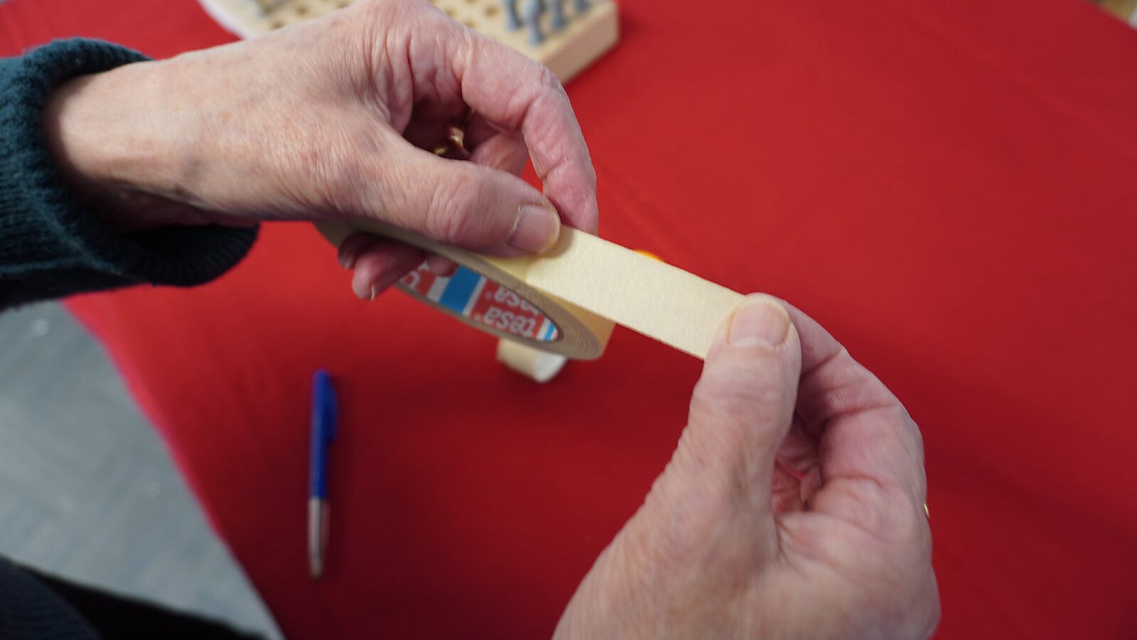 Sfincterman nr.0.0 — videostill handeling: twee handen trekken een stuk schilderstape los van de rol boven een rode tafel, met een pen en een rasterbord op de achtergrond.
