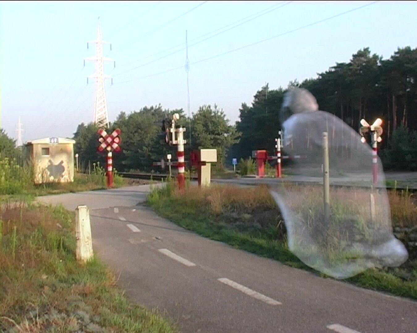 Beweging en verstilling — On the Origin of Butterflies — videostill van een landweg die naar een spoorwegovergang leidt; een halftransparante menselijke figuur beweegt door het beeld terwijl het landschap stil blijft.