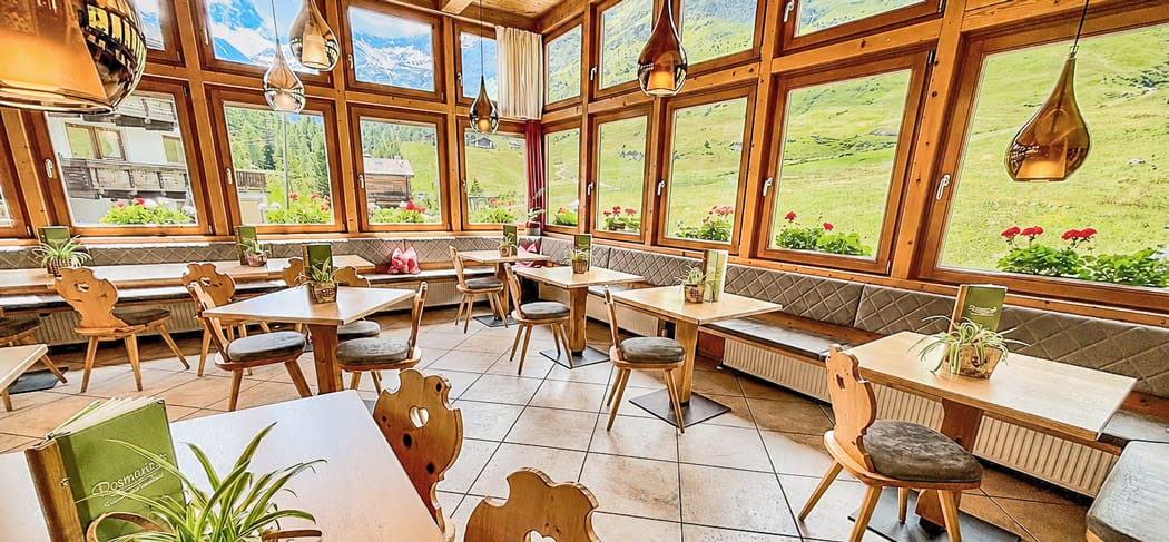 Luminoso giardino d’inverno al Ristorante Rosmarie a Plan con tavoli in legno, grandi finestre e vista sul paesaggio della Val Passiria.