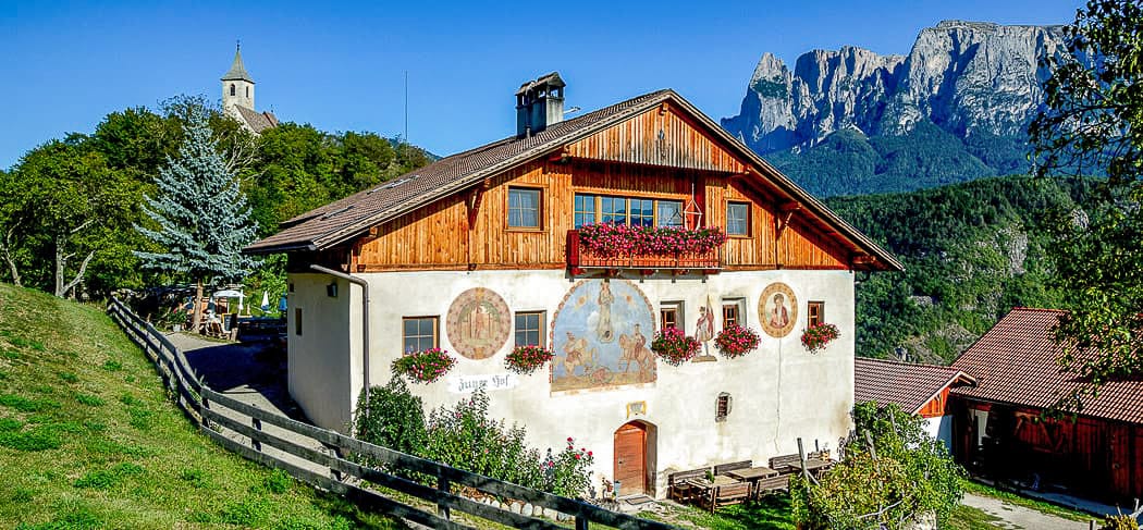 Zunerhof - Tipico maso a Longostagno sul Renon con facciata decorata, balconi fioriti e vista sulle Dolomiti in un panorama soleggiato di montagna