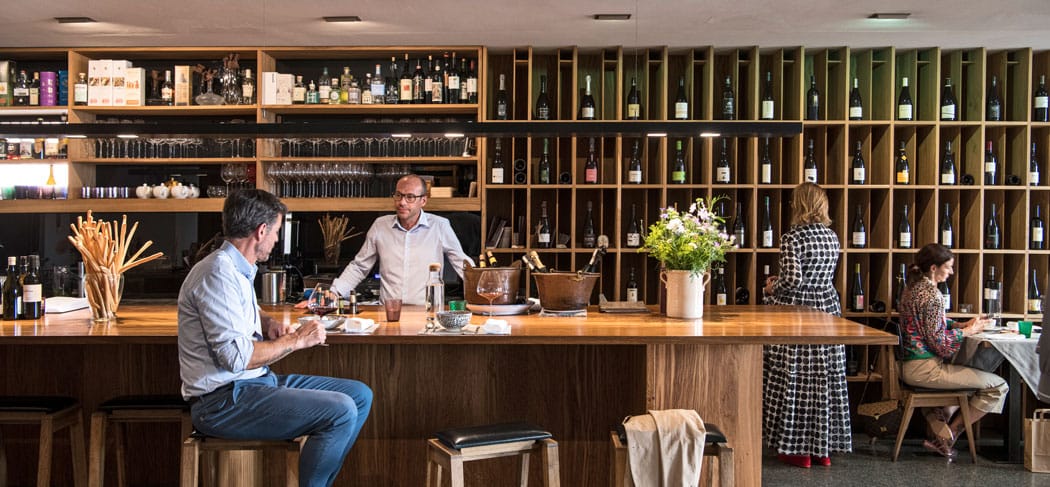Vinzenz - zum feinen Wein, – Ristorante e wine bar a Vipiteno: elegante bancone in legno, selezione di vini sulla parete, ospiti intenti a degustare vini pregiati e prelibatezze.