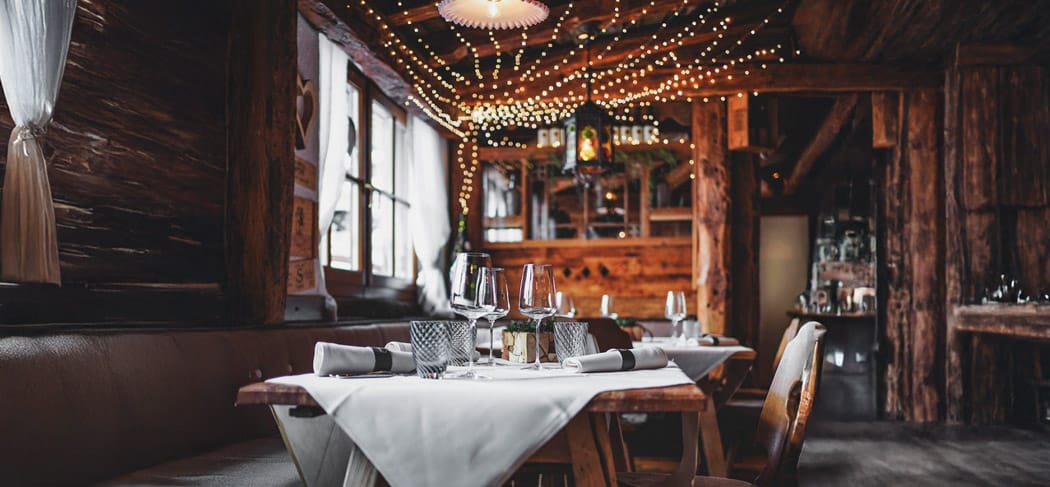 Gemütliche Stube im Restaurant Tubladel in Gröden mit rustikalen Holztischen, gedecktem Tisch, Weingläsern und stimmungsvoller Lichterkette an der Decke.