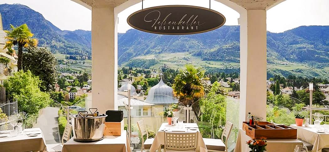 Terrasse des Restaurants Felsenkeller mit Blick über Meran, stilvoll gedeckten Tischen, Palmen und Bergpanorama – ein Logenplatz am Tappeinerweg.