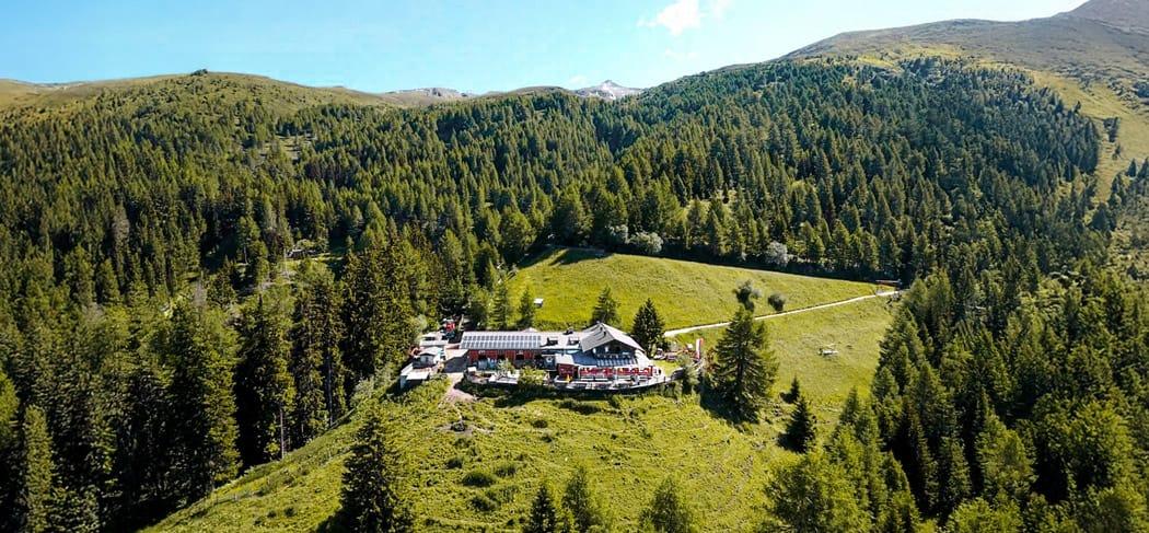 Luftaufnahme der Prantneralm oberhalb von Sterzing in Südtirol, umgeben von dichten Bergwäldern, sonnigen Wiesen und eindrucksvoller Alpenlandschaft.