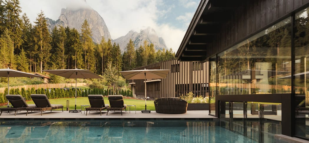 Beheizter Außenpool mit Liegen und Sonnenschirmen im Waldrast Living in Seis am Schlern, mit Blick auf Wald und die beeindruckenden Dolomiten der Seiser Alm