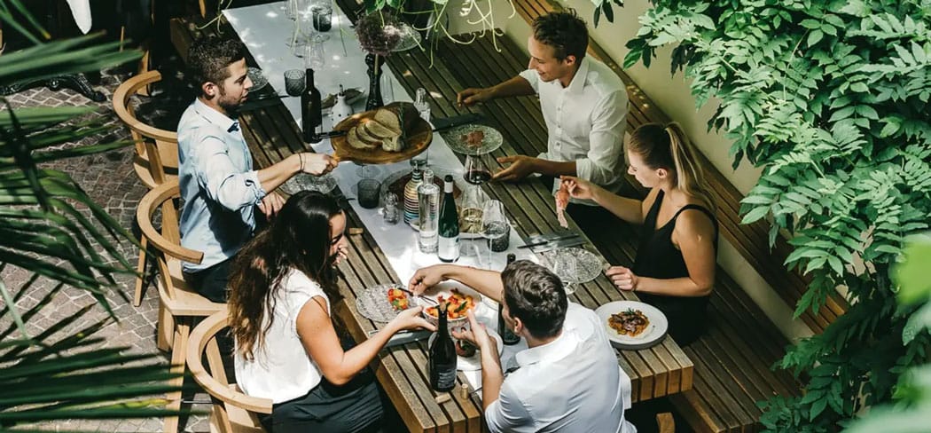 Fünf junge Erwachsene genießen gemeinsam ein stilvolles Mittagessen im schattigen Innenhof des Restaurants Vitis in Brixen.