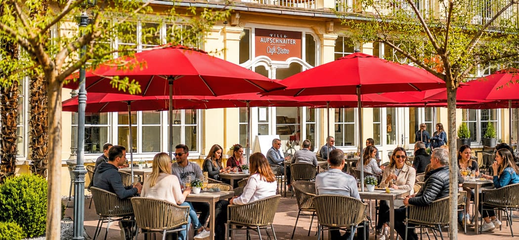 La terrazza soleggiata del Café Bistro Villa Aufschnaiter a Bolzano-Gries, con ombrelloni rossi e ospiti che fanno colazione, prendono l'aperitivo e si godono momenti di relax.