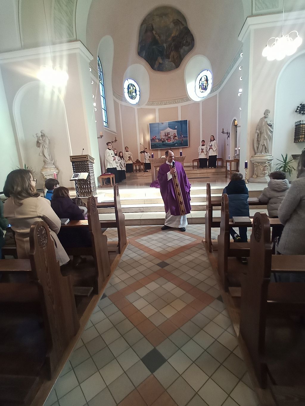 Foto zeigt einen Blick auf Pfarrer Schmit, der vor den Stufen des Altarraums steht und die Kinder befragt, die in den ersten Reihen sitzen, nach dem, wie sie sich im Gemeindeheim auf den Gottesdienst vorbereitet haben.