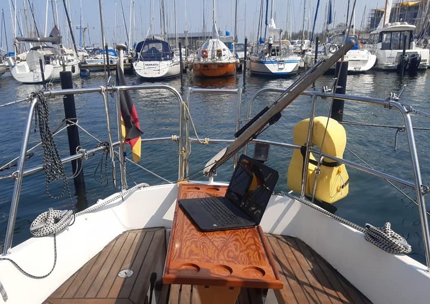 Das Heck eines Segelbootes mit Deutschlandfahne im Hafen. Auf der gegenüber liegenden Seite sind ca. 10 weitere Boote zu sehen, die im Hafen liegen. Auf einem Holztisch steht ein aufgeklappter Laptop. 