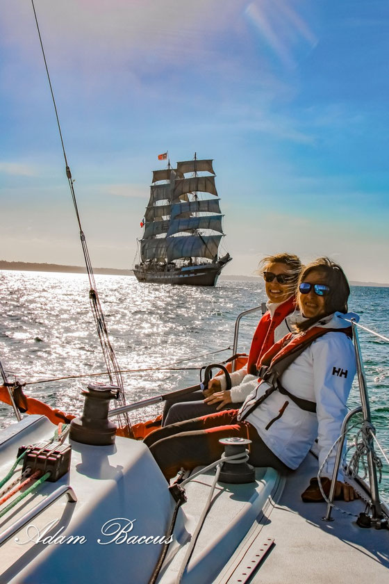 2 femmes riants sur un voilier avec Le Belem derrière elles sur ciel bleu.