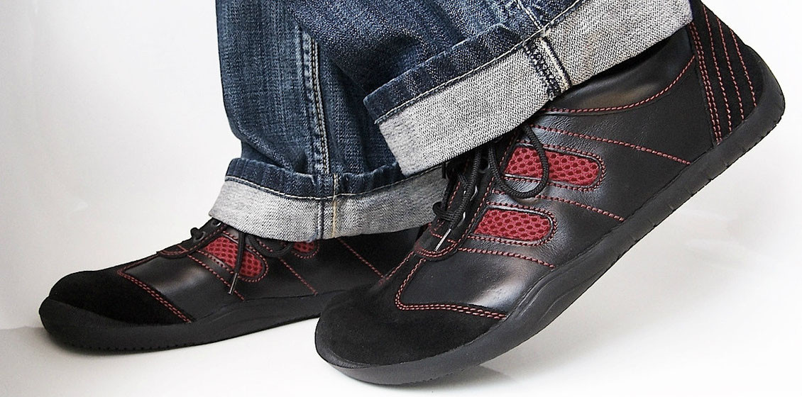 Senmotic Barfußschuhe aus schwarzem Leder mit roten Mesheinsätzen und schwarzer Sohle an den Füßen einer Frau.