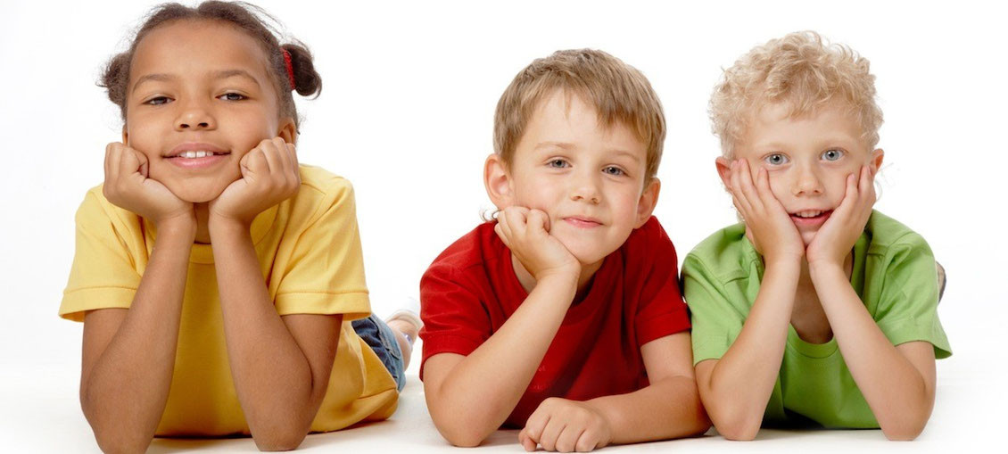 Drei Kinder zwischen 6 und 8 Jahren liegen auf dem Bauch und stützen ihren Kopf in ihre Hände und lächeln