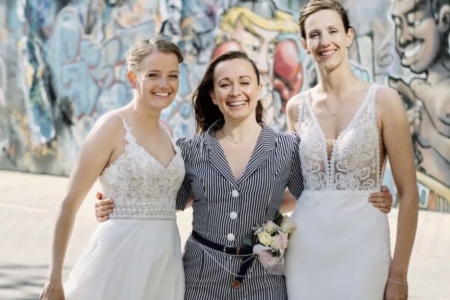 Constanze Marienfeld steht strahlend lachend zwischen ihren beiden Bräuten die beide weiße Brautkleider tragen und ebenfalls strahlen.