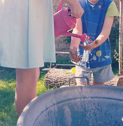 ein Kind gießt einem kleineren Kind aus der Gießkanne Wasser auf die Hände zum Waschen