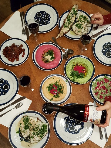 Levantische Mezze (Hummus, Babaganoush, Tahinisaucen) in bunten Farben auf einem Tisch. Dazu wird Shilo Rotwein eingeschenkt.