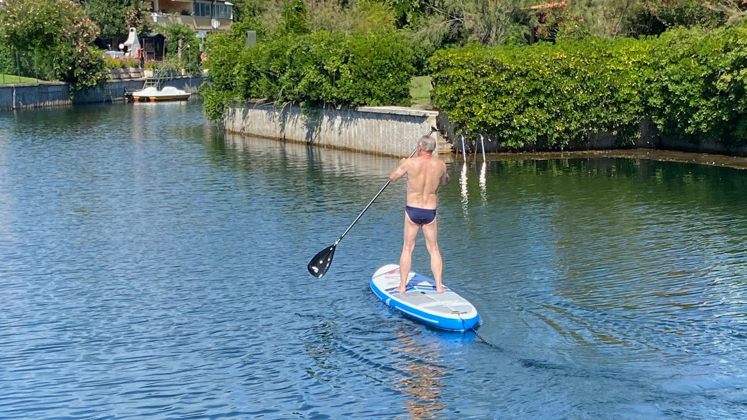 Ein Mann im Badeanzug steht auf einem Paddleboard und paddelt durch einen malerischen Fjord auf Albarella.