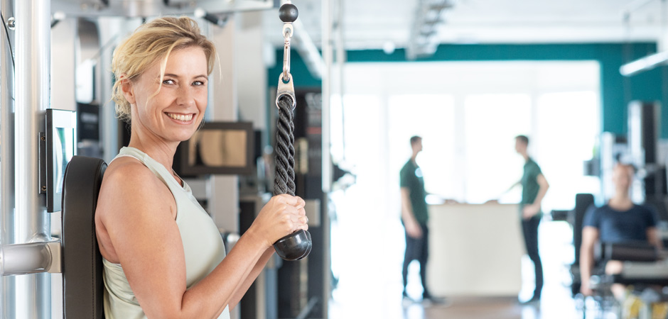 Eine Kundin beim Krafttraining. Im Hintergrund sind zwei Trainer und ein weiterer Kunde zu sehen.